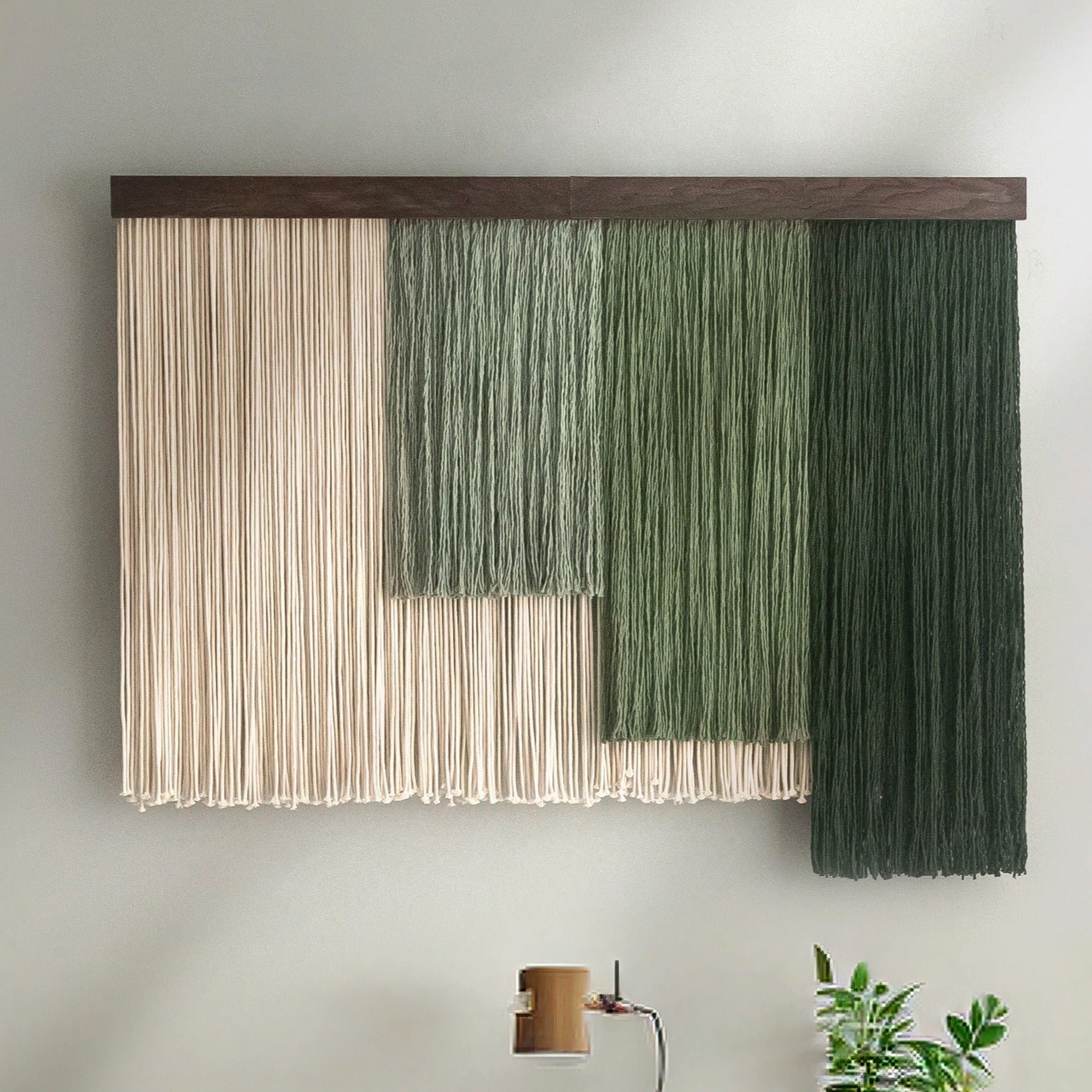 A macrame fiber wall hanging featuring various shades of green and beige cords, displayed on a wall.